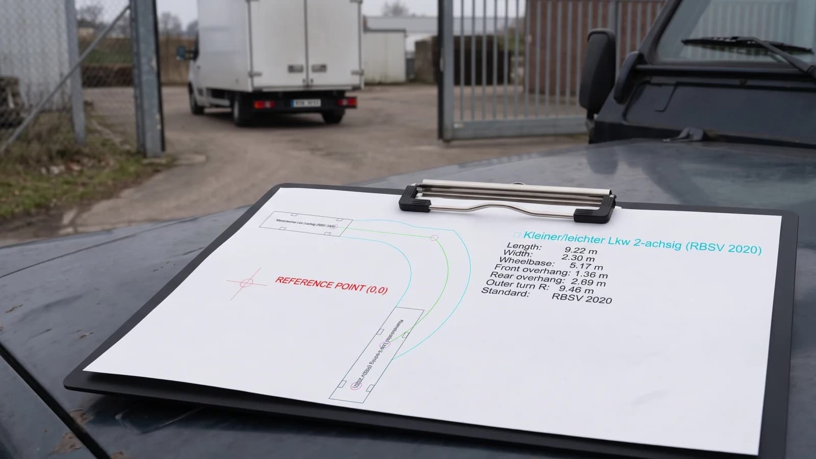 Ausgedruckte Schleppkurven-Zeichnung eines 2-achsigen LKW mit Referenzpunkt und Fahrzeug-Datenblatt auf einem Klemmbrett auf der Motorhaube, im Hintergrund Hoftor und LKW
